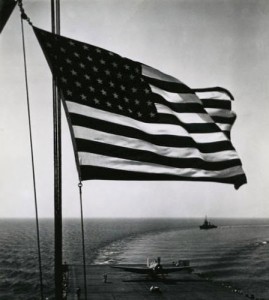 An American flag flies from an aircraft carrier as a plane lands. (US Naval Academy)