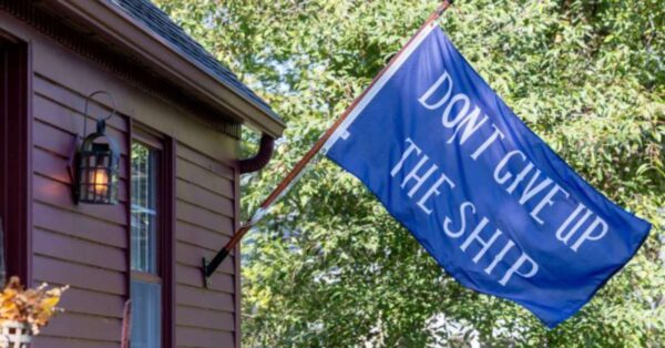 blue 'don't give up the ship' flag hanging from a pole mounted to a small bown house in front of verdant woods
